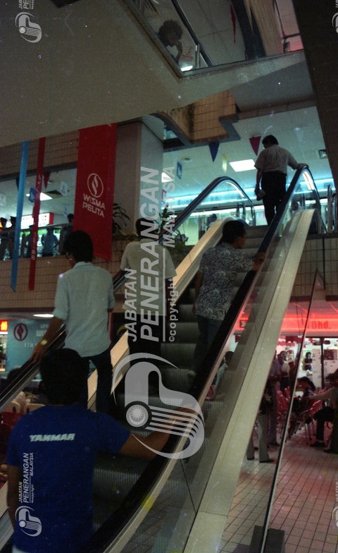 Then: Shot of people going up the escalator in the previous image (five at once). The backdrop of the stage mentioned
				earlier is visible. The planters and banners are also visible.