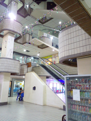 Now: The space is now lit with bright floodlights mounted to the columns. The banners and plants are gone. Fairy lights cross the first floor void.
				An extra room has been built beneath the single escalator visible.
				No people are visible except one and a half coffee shop patreons, blocked by the escalator room.