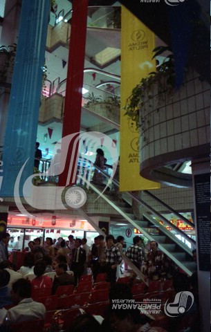 Then: Inside the single enclosed plaza of the building's shopping centre. There's a sizable crowd. Red, yellow, and blue banners are visible 
				decending from the top beyond the frame, stopping just below the first floor (British floor numbering, Floor 1 is the first floor above the ground).
				Red plastic seats, many occupied, face towards a stage out of frame. It is reflected in the mirrored pannels on the 
				escalator, revealing it has a grey backdrop. A few planters are visible. 
				They are all built into the columns and  while not exactly overflowing, are still filled.