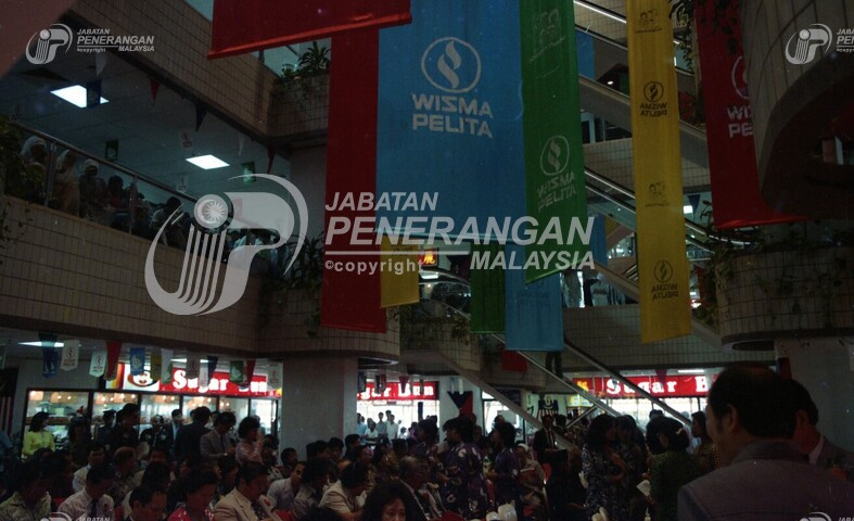 Then: Facing across the central plaza shows a large crowd of people seated, facing the yet elusive stage. The planters and banners
				mentioned earlier are visible. There are at least a dozen, and come in white on red, white on green, 
				white on blue, and black on yellow.
				This image faces the opposite way from the last ones, and faces a shop with a sign reading "Sugar Bun".
				There is no "now" image.