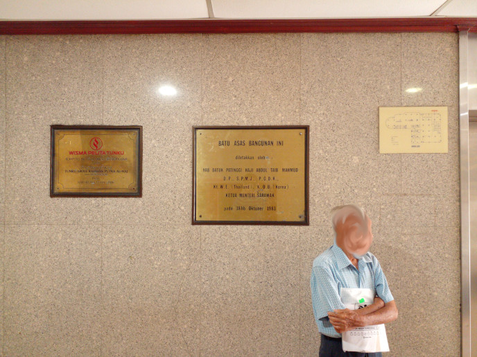 two plaques and one sign visible in a granite-clad smoke lobby.
			left to right:
			Announce opening date ((medium size, 1986), groundstone laid date (biggest, 1983), 
			and a ground-floor fire plan (smallest).
			A grampa stands to the right of the second plaque.
