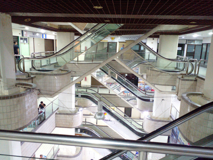 Now: So much for subtle lighting. The downlighters have been turned off, 
				replaced by the floodlights mentioned earlier, on the second floor, 
				and seemingly the superior brightness of LED lighting tech. The mezzanine escalators are not in use at all and in fact were not running.
				One downlighter dangles out of the ceiling lattice.