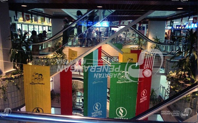 Then: On the fourth floor of the shopping centre. The banners are hung from cables strung across this floor. 
				The ceiling is visible: it consists of a dark wood lattice inset with downlighters. In the center is a hole through
				which a pair of crossed escalators rise into, leading into a mezzanine. The escalators are in busy use.