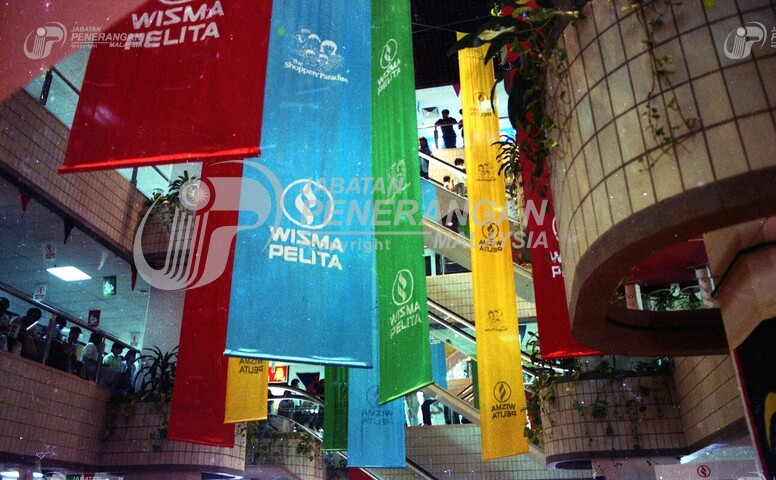 Then: Facing upwards shows the banners in more detail and a bit of the darkened ceiling of the plaza.
					The banners all read "Wisma Pelita - The Shoppers' Paradise". There is no "now" image.